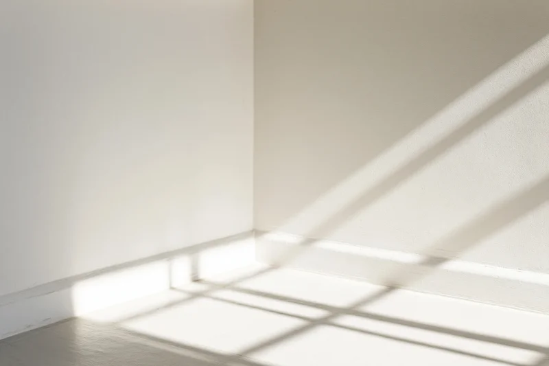 Diagonal window frame shadows cast across a bare white interior corner