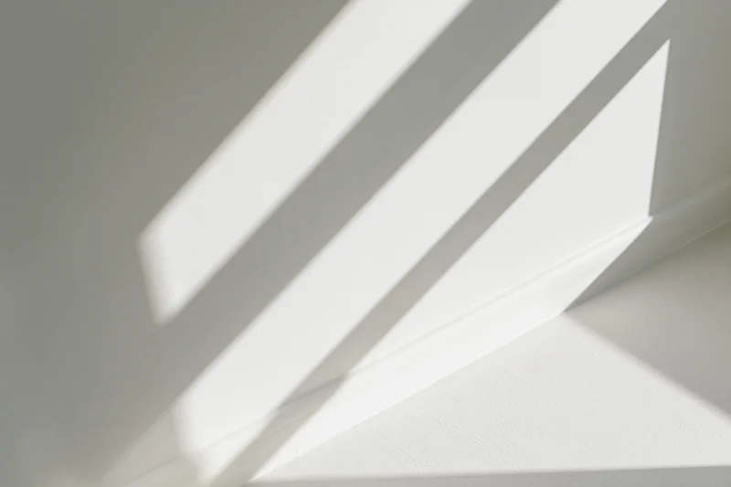 Diagonal shadows from a window frame cast across a white interior wall and ceiling