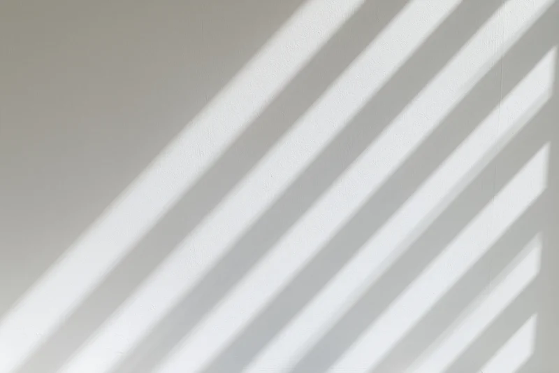 Diagonal bands of light and shadow from a window falling across a white wall indoors