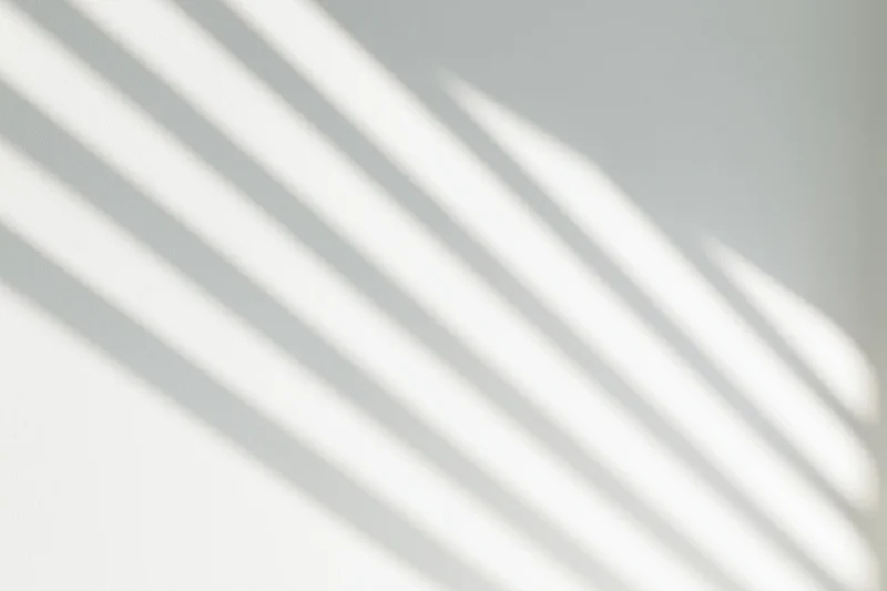 Diagonal shadow lines from a window cast across a plain white wall in soft natural light