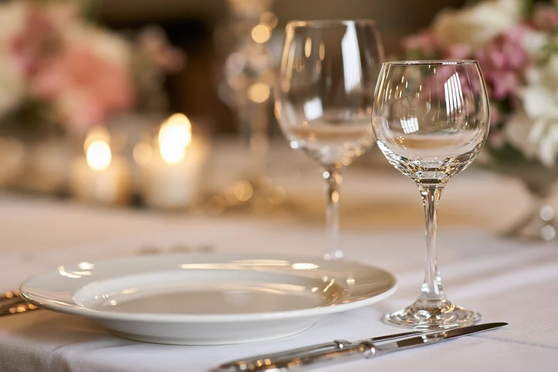Elegant dinner table setting with crystal wine glasses and white plate on linen tablecloth, candles and floral centerpiece in background