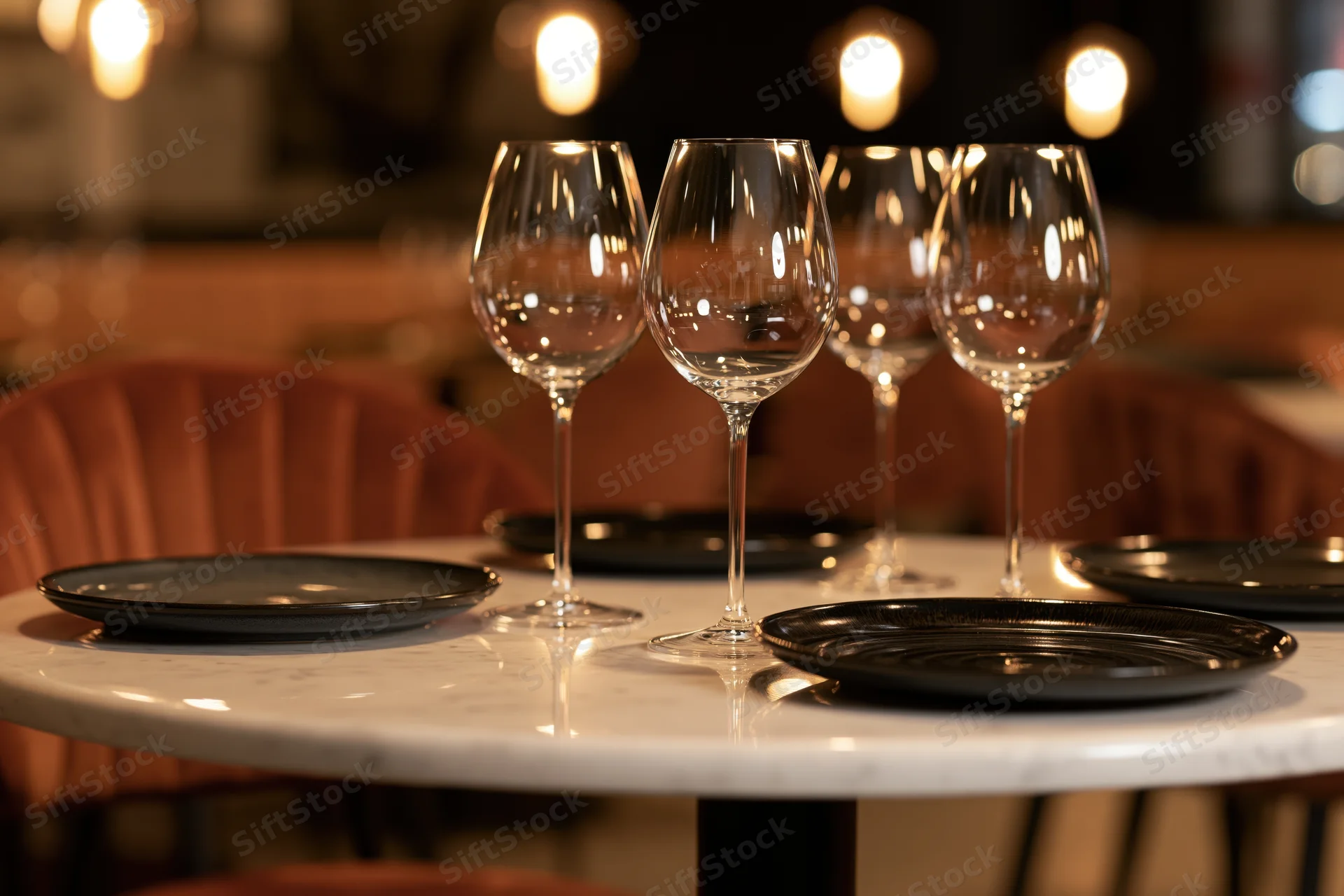 Four empty wine glasses and black plates arranged on a marble table in a dimly lit restaurant interior