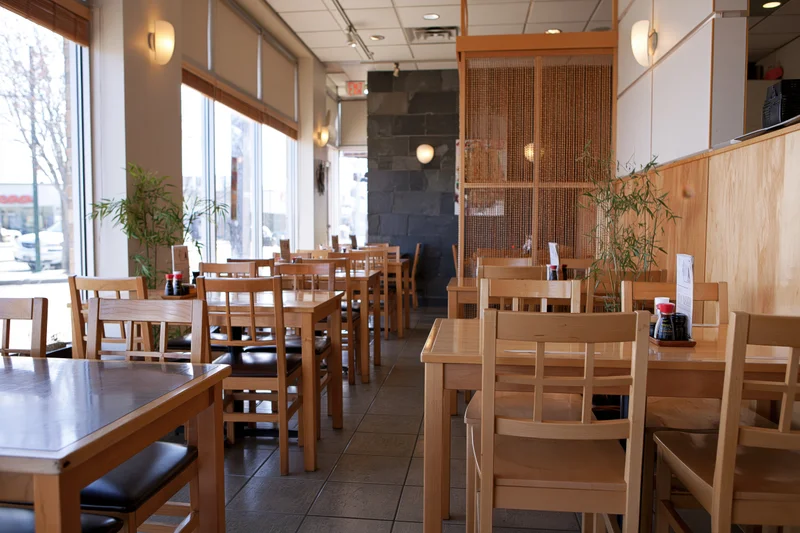 Empty wooden tables and chairs arranged in rows inside a casual japanese restaurant dining room with large windows and bamboo partitions