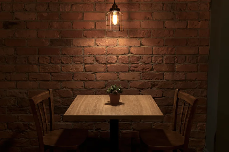 Square wooden table with two chairs and a potted plant under a pendant lamp in a brick-walled cafe corner