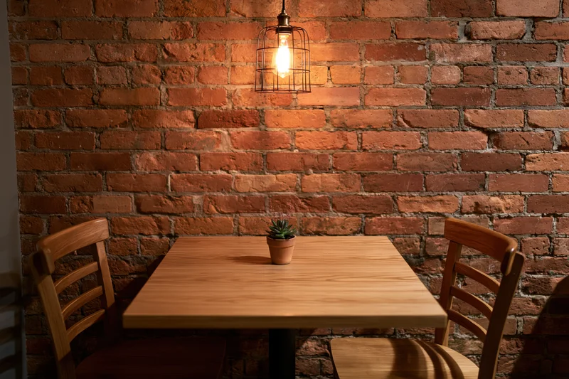 Square wooden table with small potted succulent between two chairs against exposed brick wall under warm pendant light in cafe corner