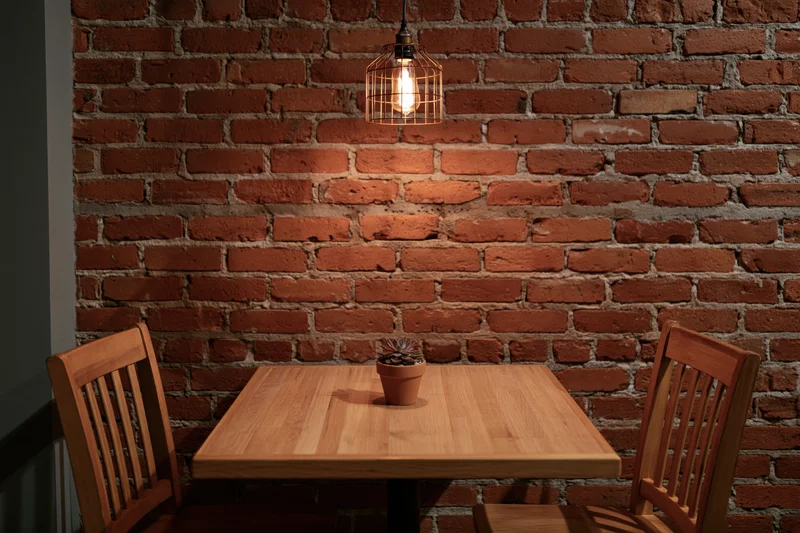 Square wooden table with small potted succulent between two chairs against exposed brick wall under warm pendant light