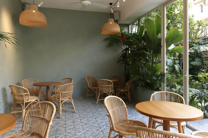 Empty cafe interior with rattan chairs and round wooden tables arranged near large glass doors overlooking greenery