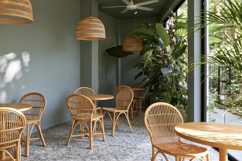 Empty cafe interior with rattan chairs and round wooden tables near large glass doors and indoor plants