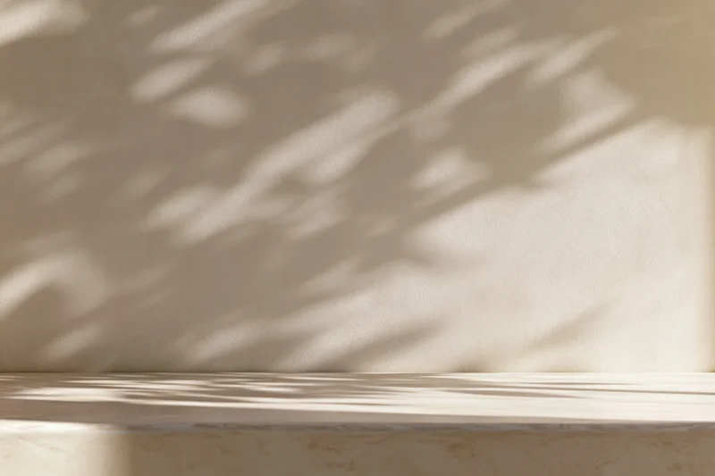 Smooth cream plaster wall with dappled sunlight shadows above a light wooden tabletop surface