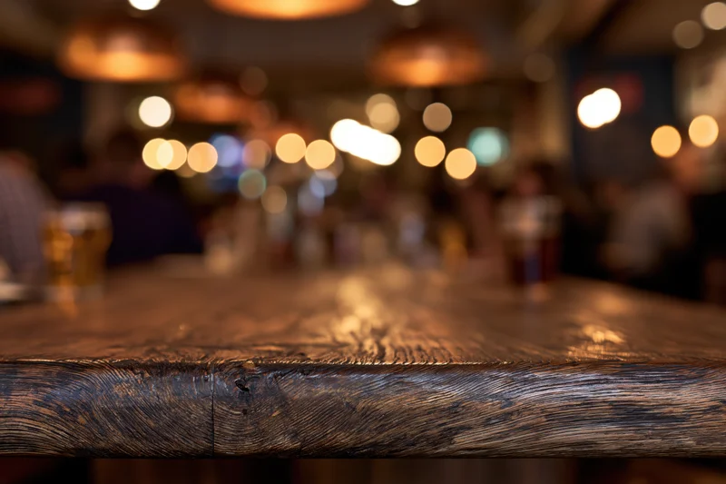 Dark wooden bar counter in sharp focus with blurred background of patrons and warm lights in restaurant interior