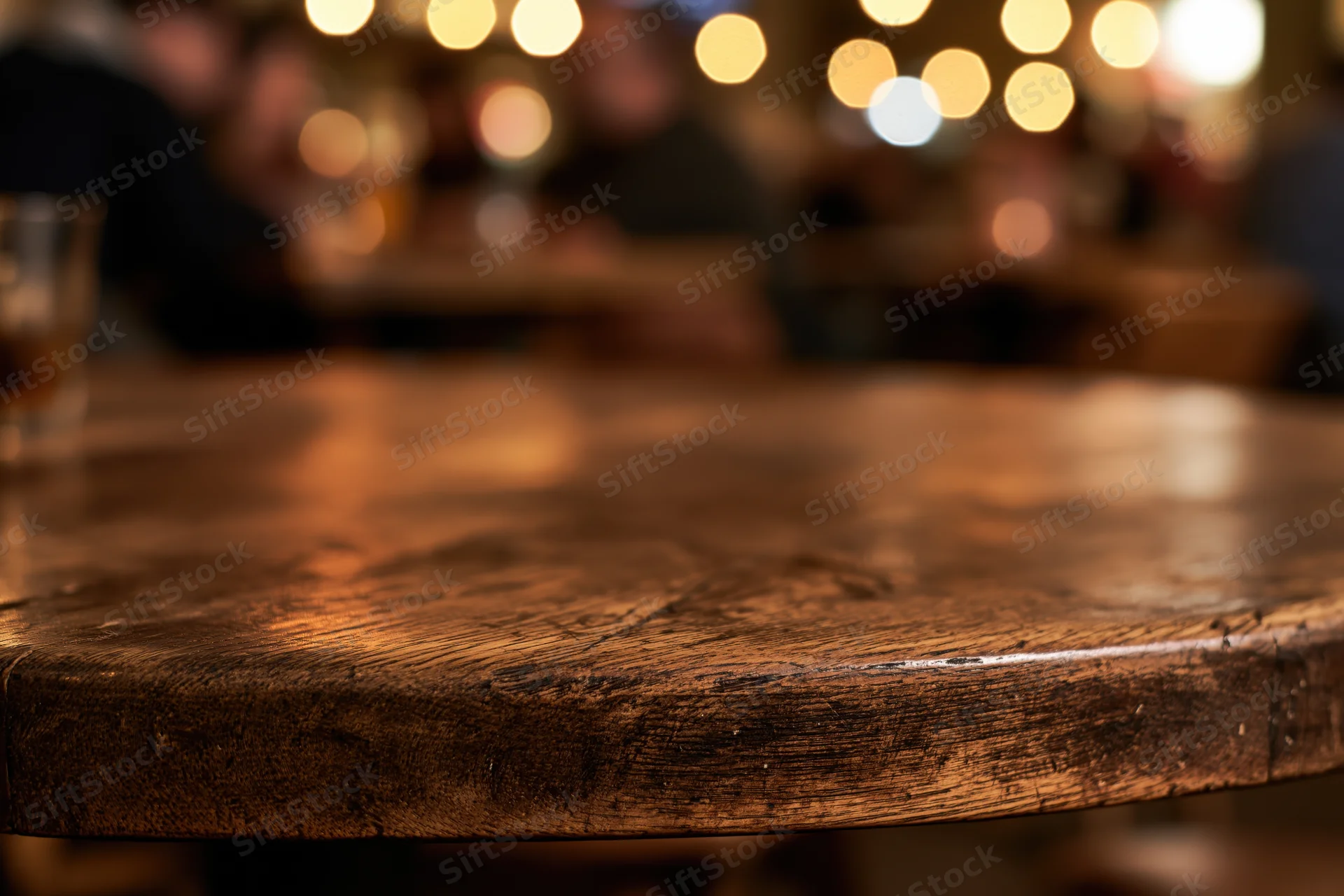 Closeup of dark wooden table surface in a dimly lit bar with blurred patrons and warm bokeh lights in background