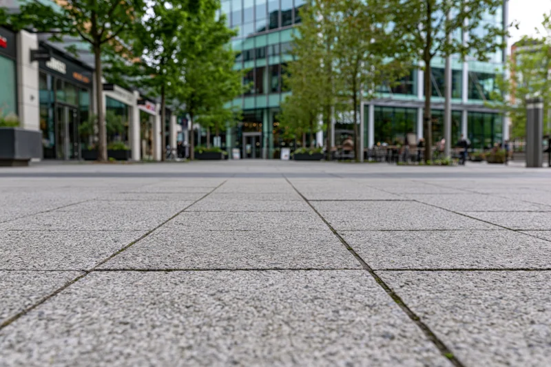 Low-angle view of gray stone pavement in a modern urban plaza with glass buildings and trees in the background