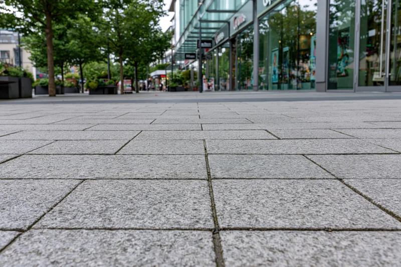 Low-angle view of gray stone pavement tiles leading toward modern glass storefronts in an urban plaza