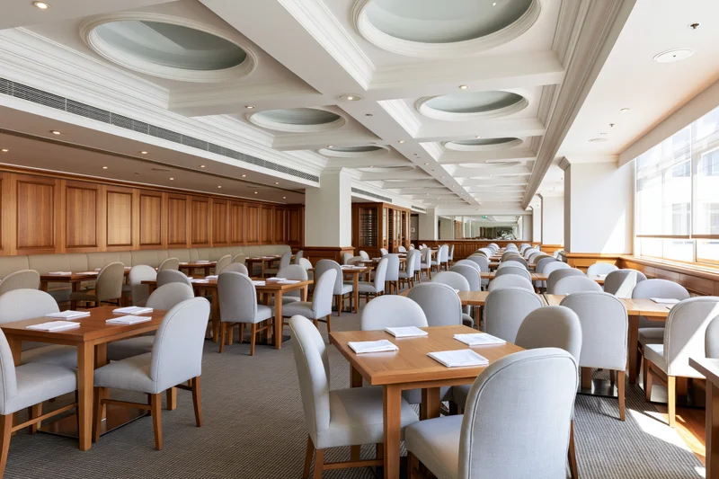 Empty hotel restaurant dining room with rows of rectangular wooden tables and white chairs under coffered ceiling with recessed lights