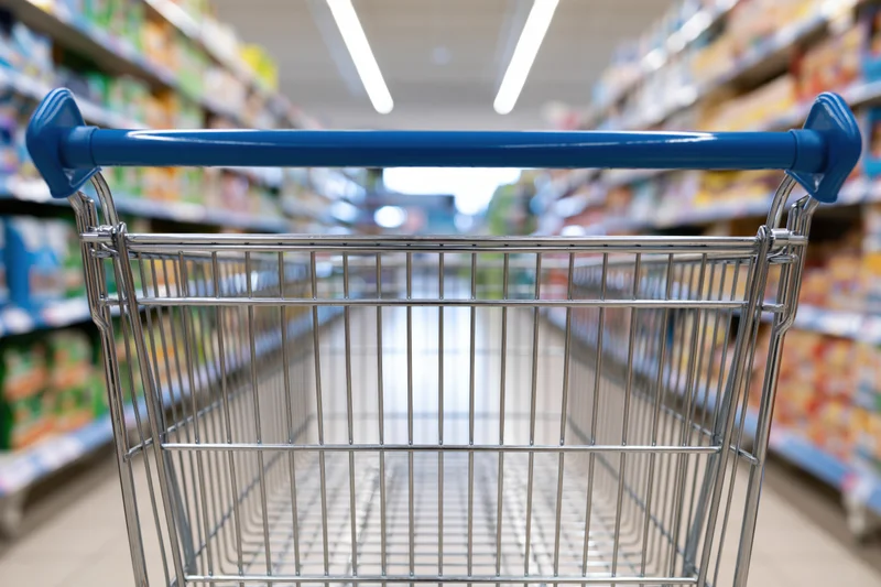 Empty metal shopping cart with blue plastic handles viewed down a supermarket aisle
