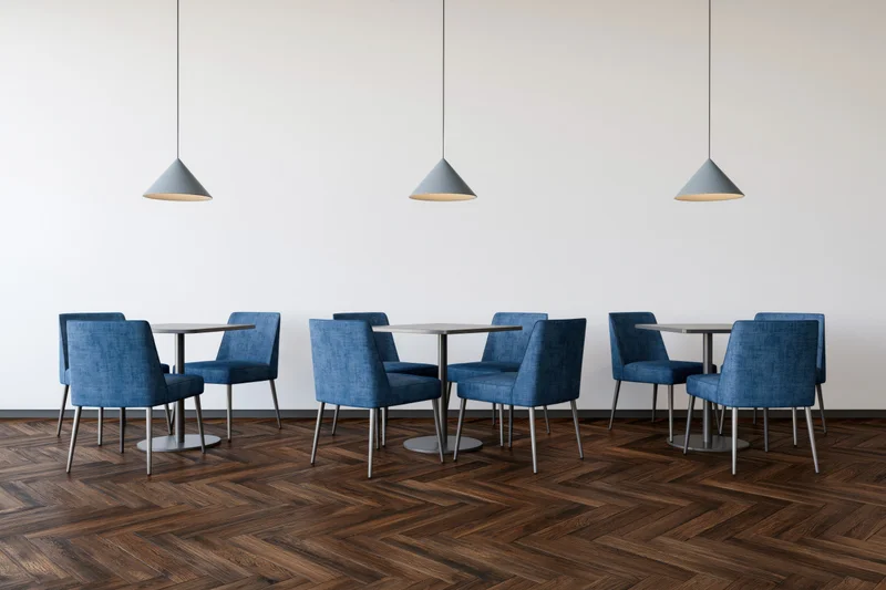 Modern cafe interior with small square tables and blue chairs arranged in rows on herringbone wood flooring