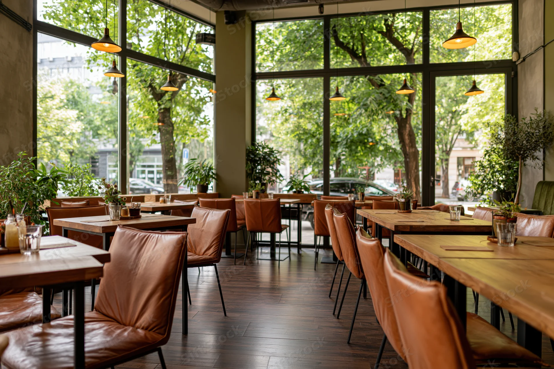 Modern cafe interior with wooden tables and brown leather chairs arranged near large windows overlooking green trees and city street