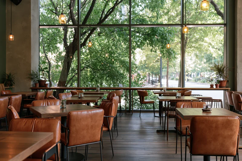Empty modern cafe interior with wooden tables and brown leather chairs by large windows overlooking trees