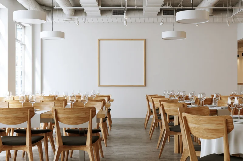 Empty modern restaurant dining room with rows of wooden chairs and tables set with wine glasses and white tablecloths under pendant lights and exposed ceiling pipes