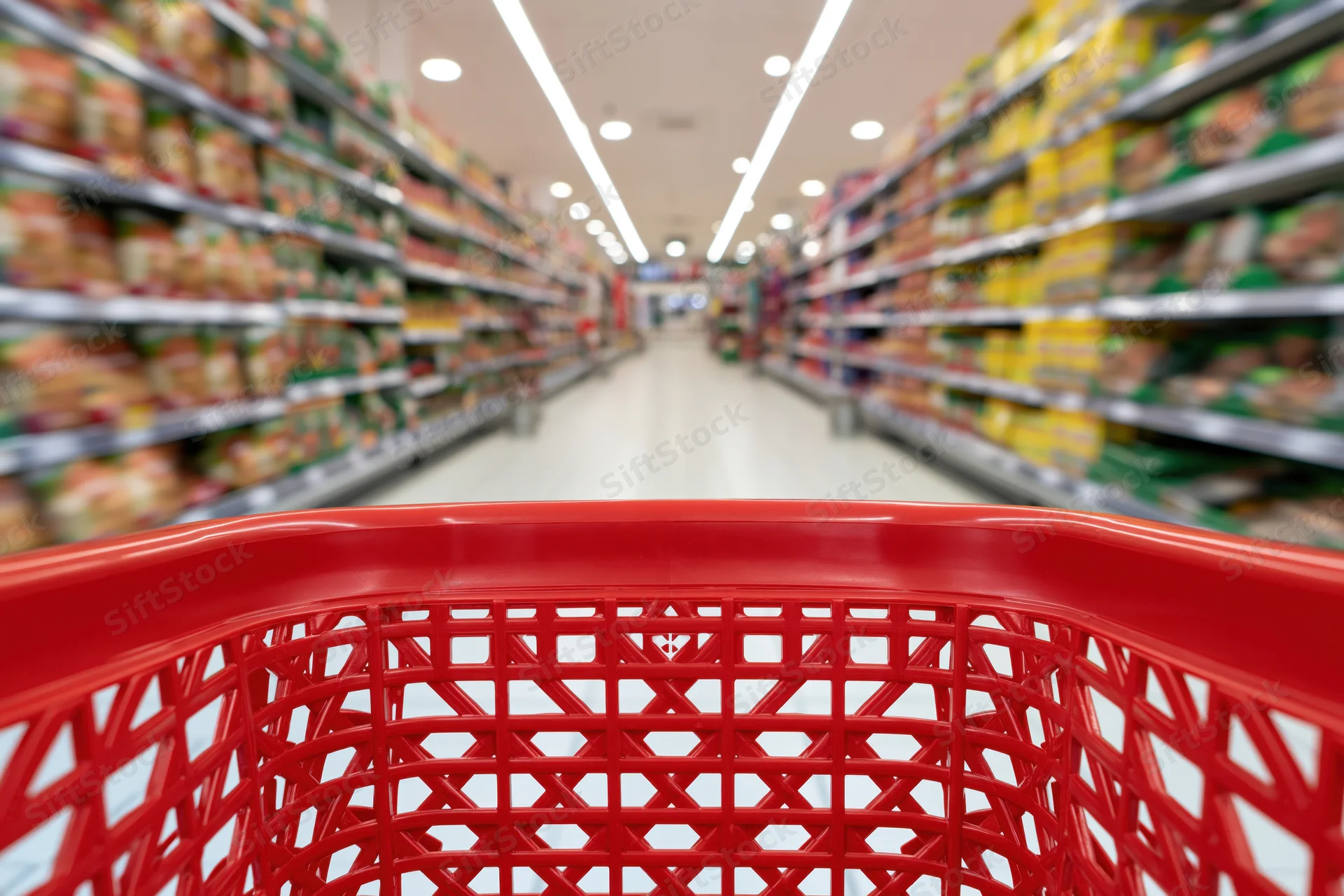 Red plastic shopping basket viewed from inside looking down supermarket aisle with shelves of products on both sides