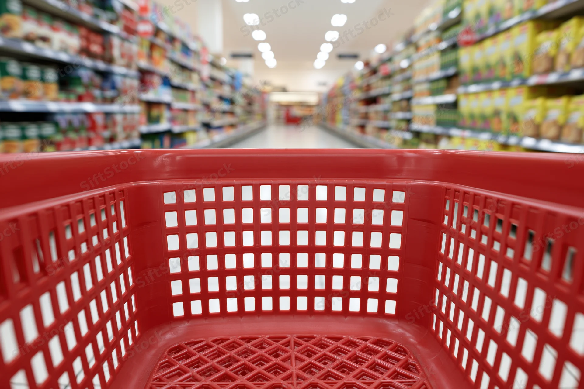 Empty red plastic shopping basket viewed from shopper's perspective down supermarket aisle