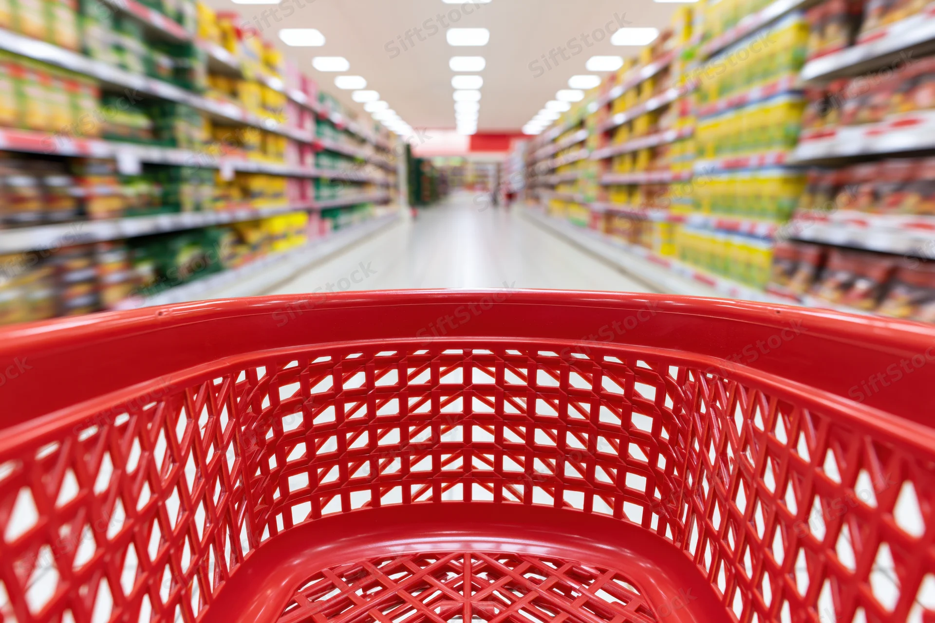 Red plastic shopping basket viewed from inside looking down supermarket aisle