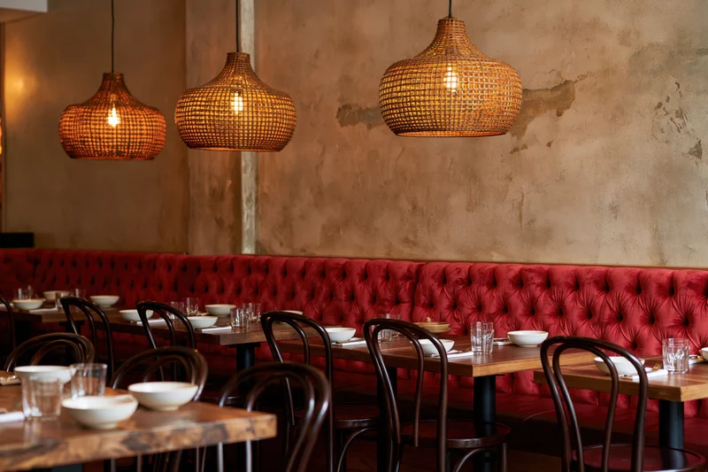 Empty restaurant dining room with wooden tables and red tufted booths under wicker pendant lights