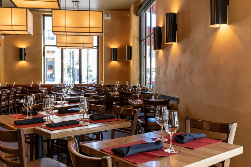 Empty restaurant dining room with wooden tables set for service, warm lighting, and large windows overlooking street