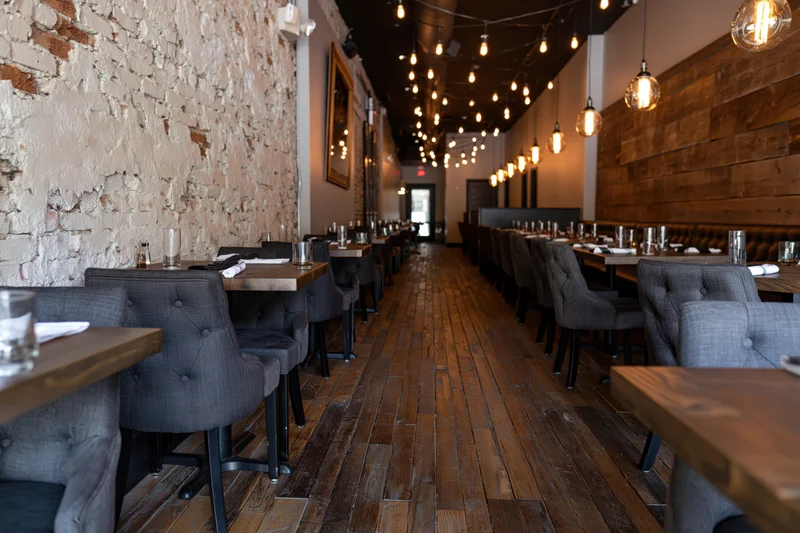 Empty restaurant interior with rows of dark tufted upholstered chairs and wooden tables along a brick wall and hardwood floor