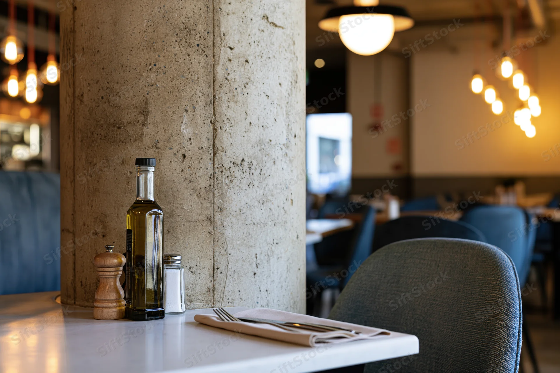 Empty restaurant table set with olive oil bottle, salt, pepper grinder and napkin with cutlery, near concrete pillar and chairs under warm pendant lights