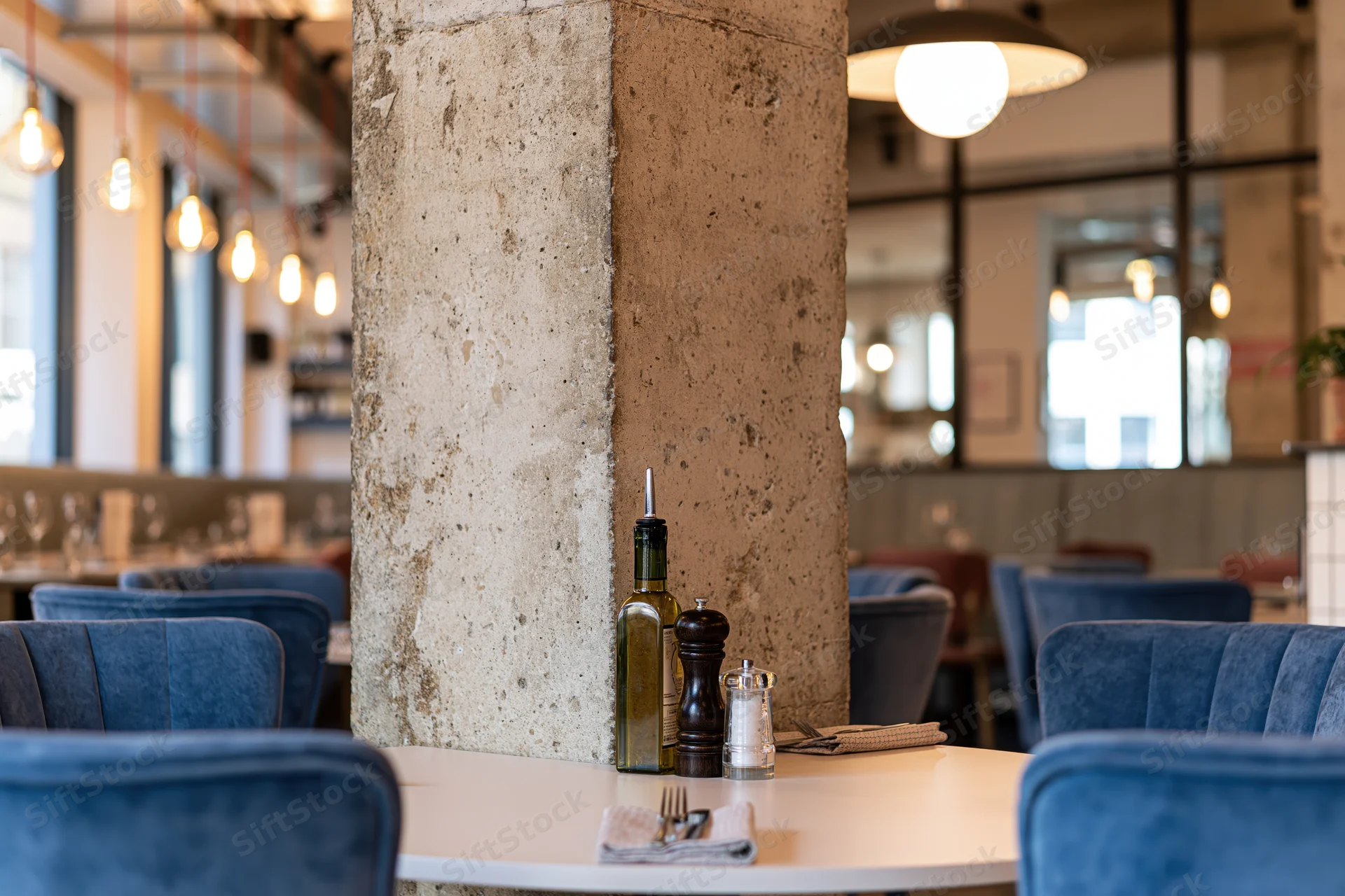 Round white table set with olive oil bottle, pepper shaker, salt shaker, napkin and cutlery in modern restaurant interior with blue velvet chairs and concrete pillar