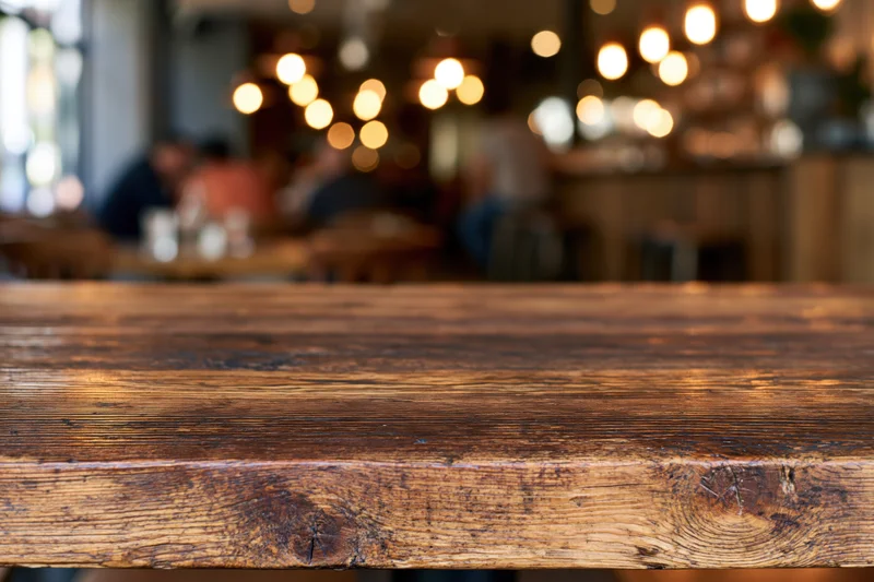 Rustic wooden table surface in sharp focus with blurred restaurant interior and warm lighting in background