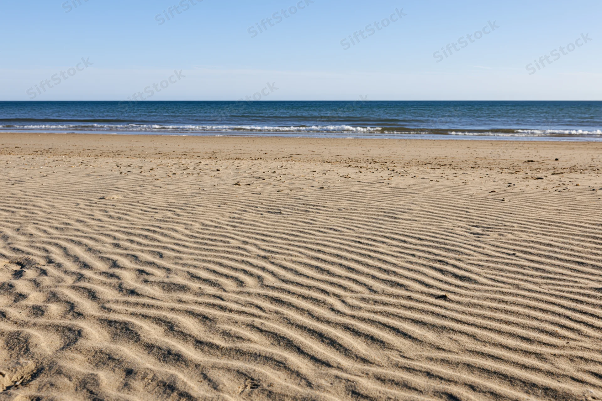 Sandy beach with rippled patterns stretching to calm ocean under clear blue sky