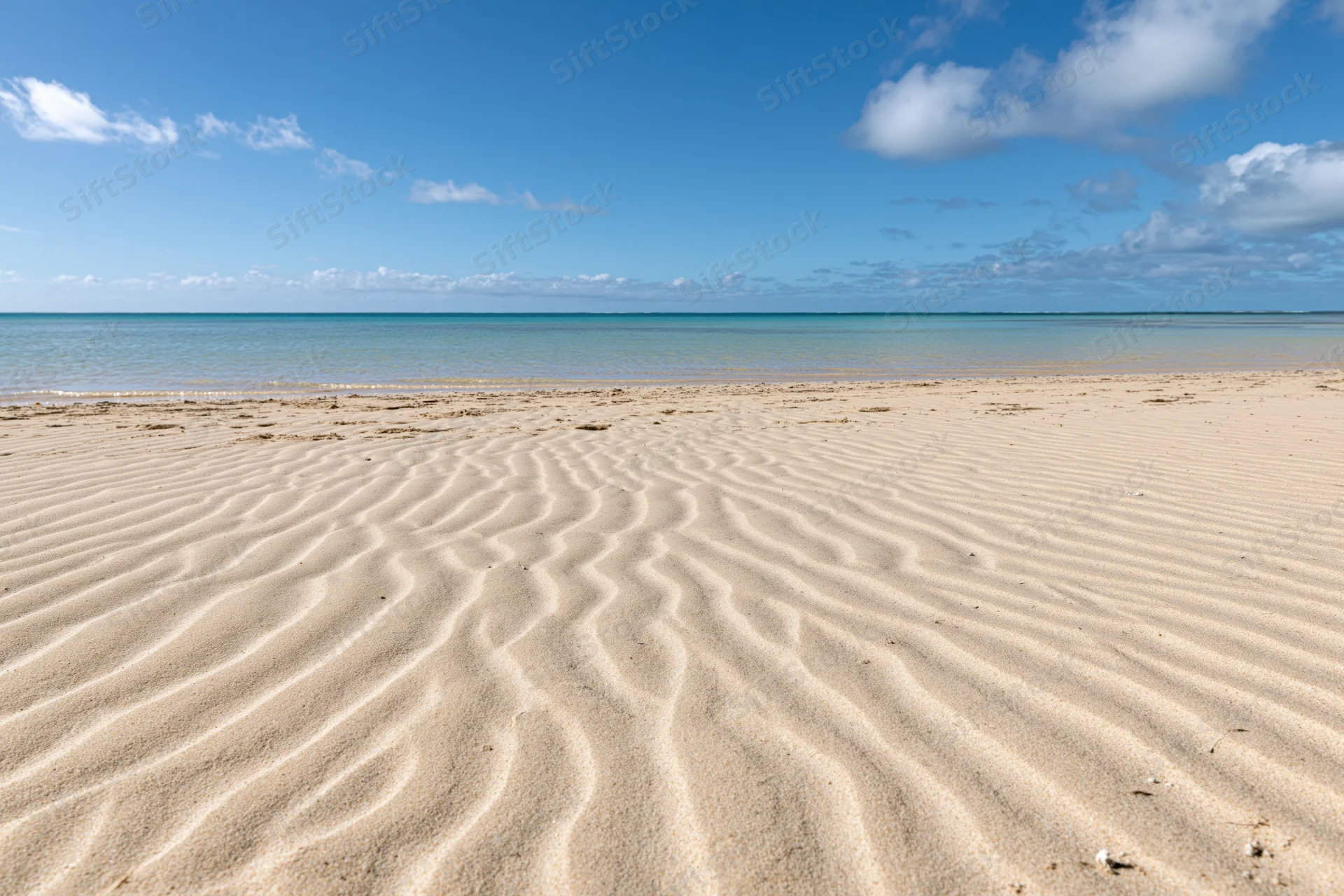 Rippled sand on a tropical beach under a clear blue sky with scattered clouds