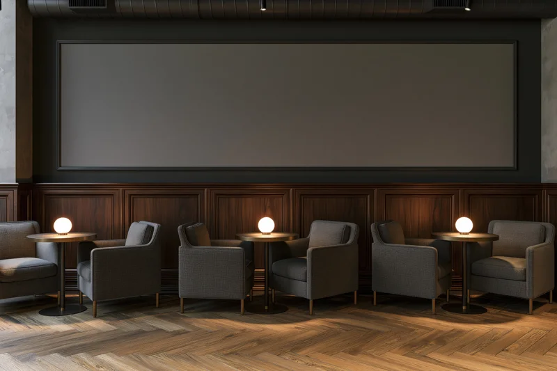 Empty upscale lounge with gray armchairs and round tables arranged in front of a large screen under warm lighting