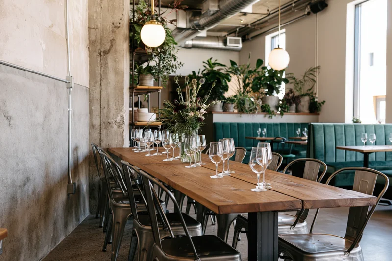 Long wooden dining table set with wine glasses and floral arrangement in modern industrial restaurant interior with exposed pipes and greenery