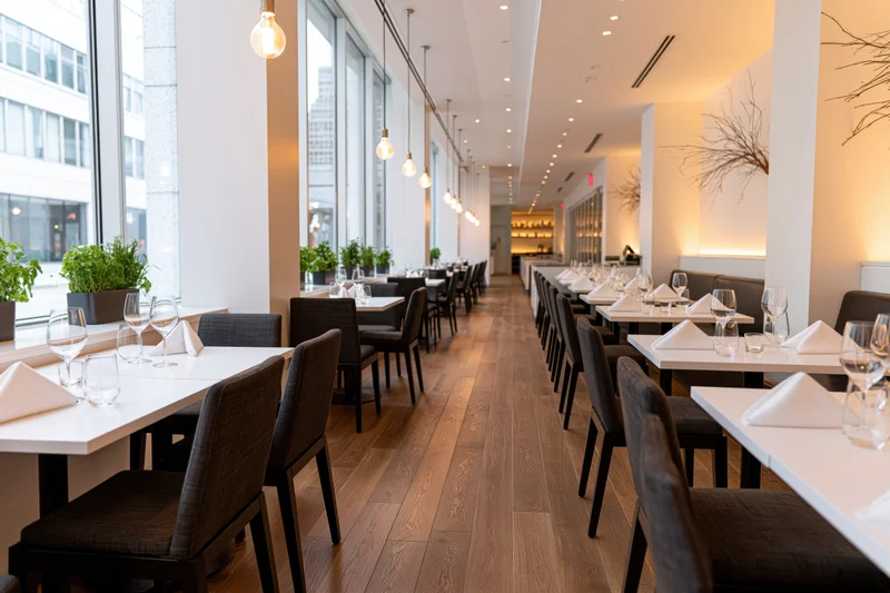 Empty upscale restaurant dining room with tables set for service along a wooden floor near large windows