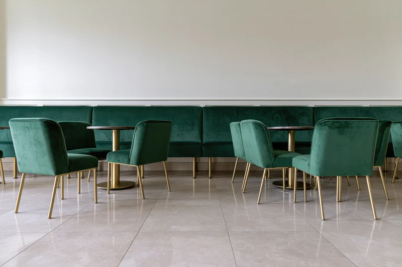 Empty upscale restaurant interior with emerald velvet chairs and gold legs arranged along a white wall with marble flooring