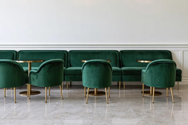 Row of emerald velvet chairs and sofas with gold legs and round tables in an upscale restaurant interior with white walls and marble floor