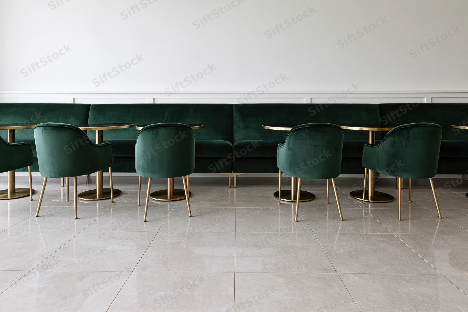 Row of emerald velvet chairs and gold-legged tables along a white wainscoted wall in a modern restaurant interior