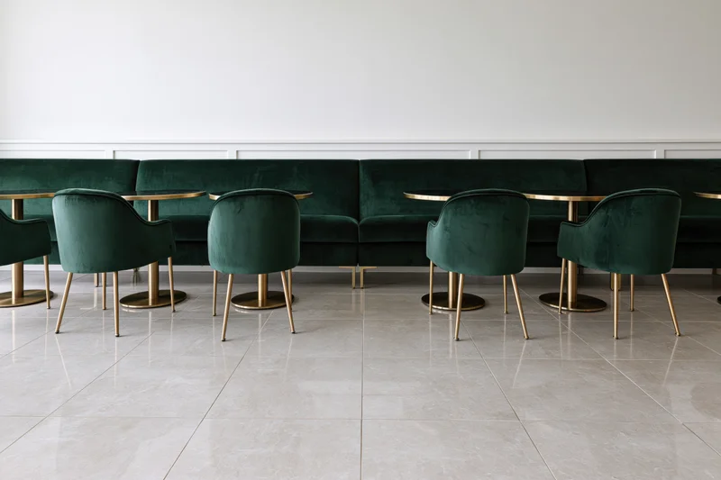 Row of emerald velvet chairs and gold-legged tables along a white wainscoted wall in a modern restaurant interior