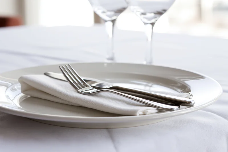 White ceramic dinner plate with folded linen napkin and silverware on a tablecloth, set for dining