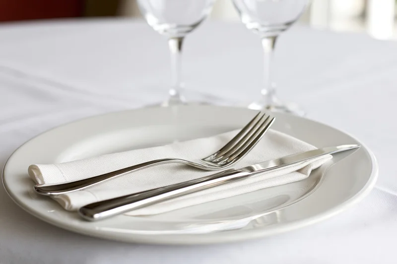 White dinner plate with folded linen napkin and silverware on a tablecloth, two wine glasses in background