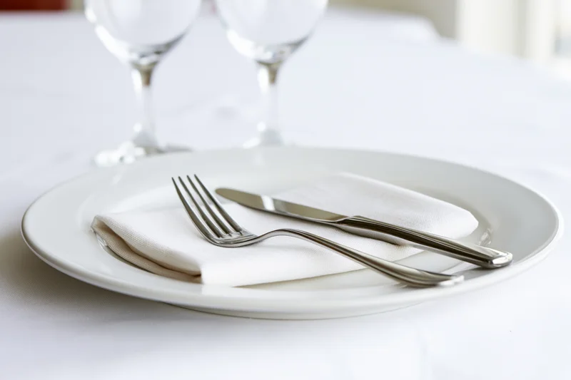 White dinner plate with folded linen napkin and silverware on a tablecloth, two wine glasses in background