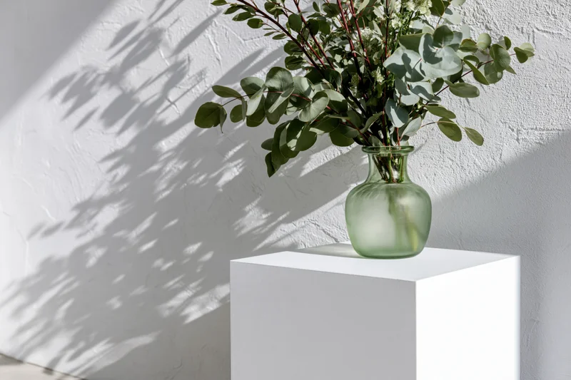 Green glass vase with eucalyptus branches on white pedestal against textured wall
