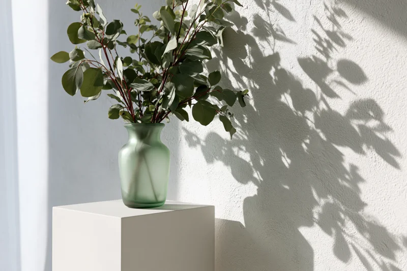 Green glass vase with eucalyptus branches on white pedestal casting leaf shadows on textured wall