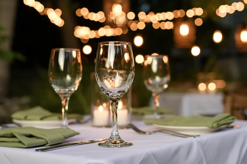 White-clothed table set with wine glasses and green napkins under string lights at night