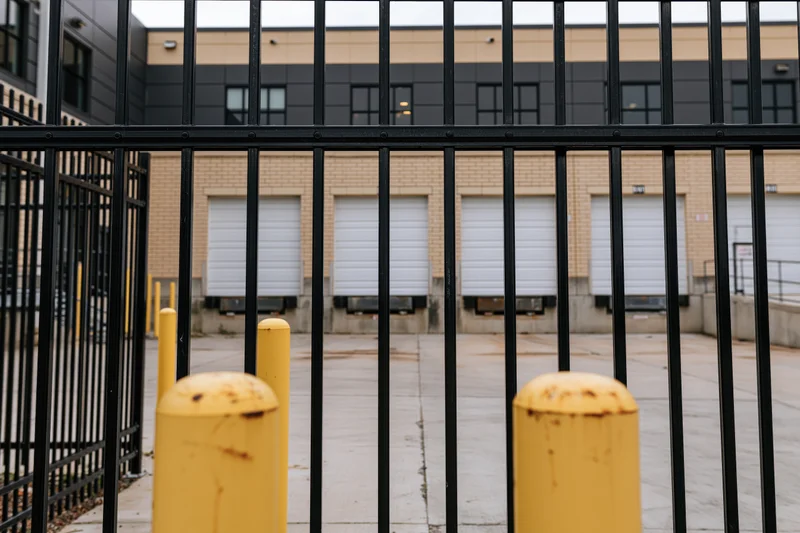 View through black metal fence of warehouse loading dock with white roll-up doors and yellow bollards on concrete pavement