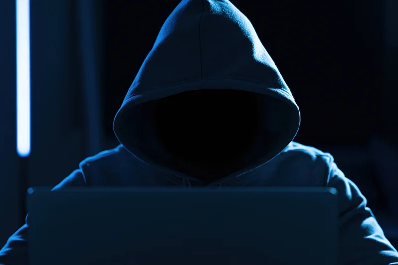 Hooded figure working on laptop in dark blue-lit room