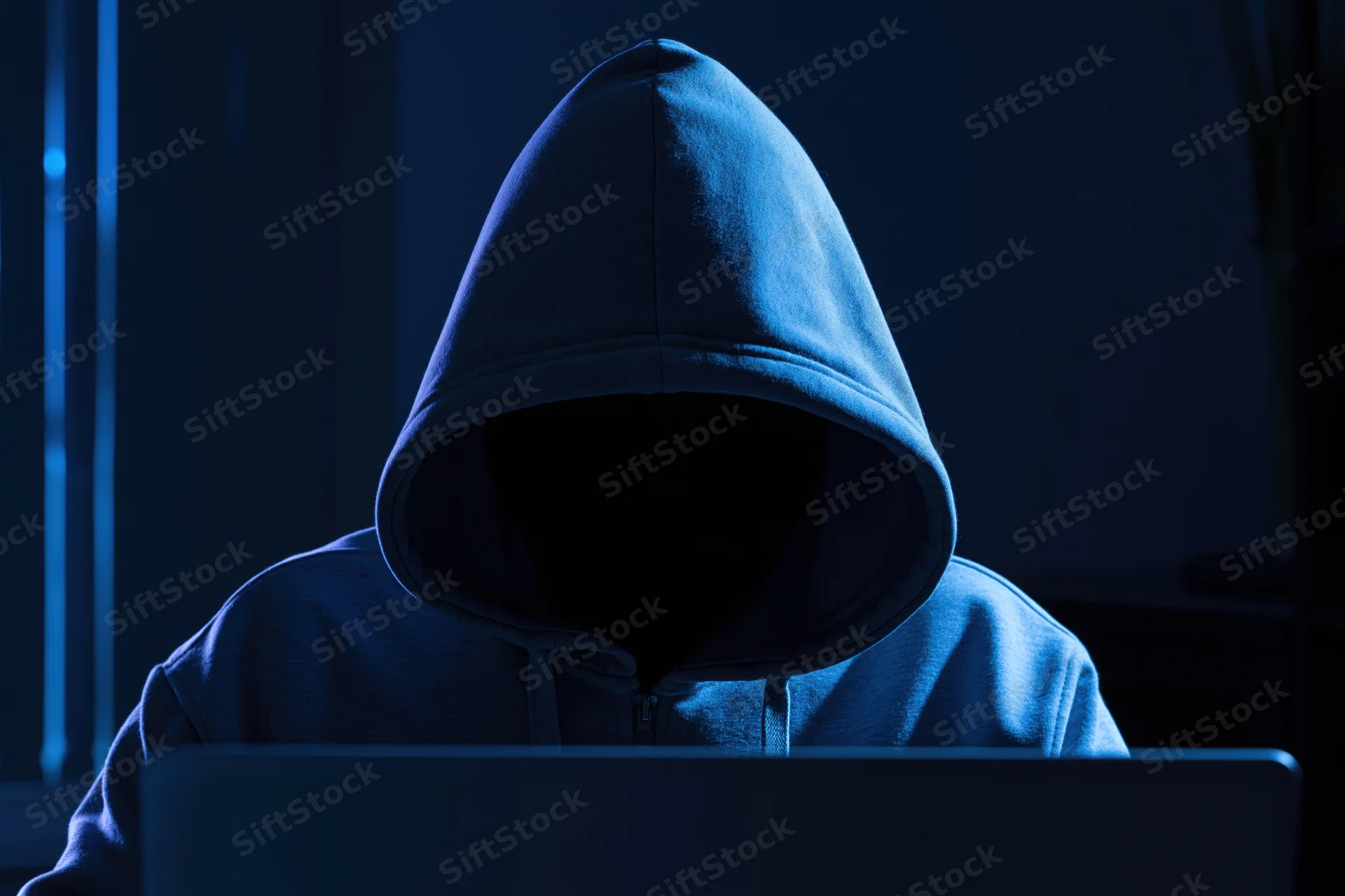 Hooded figure sitting behind laptop in dark room with blue lighting